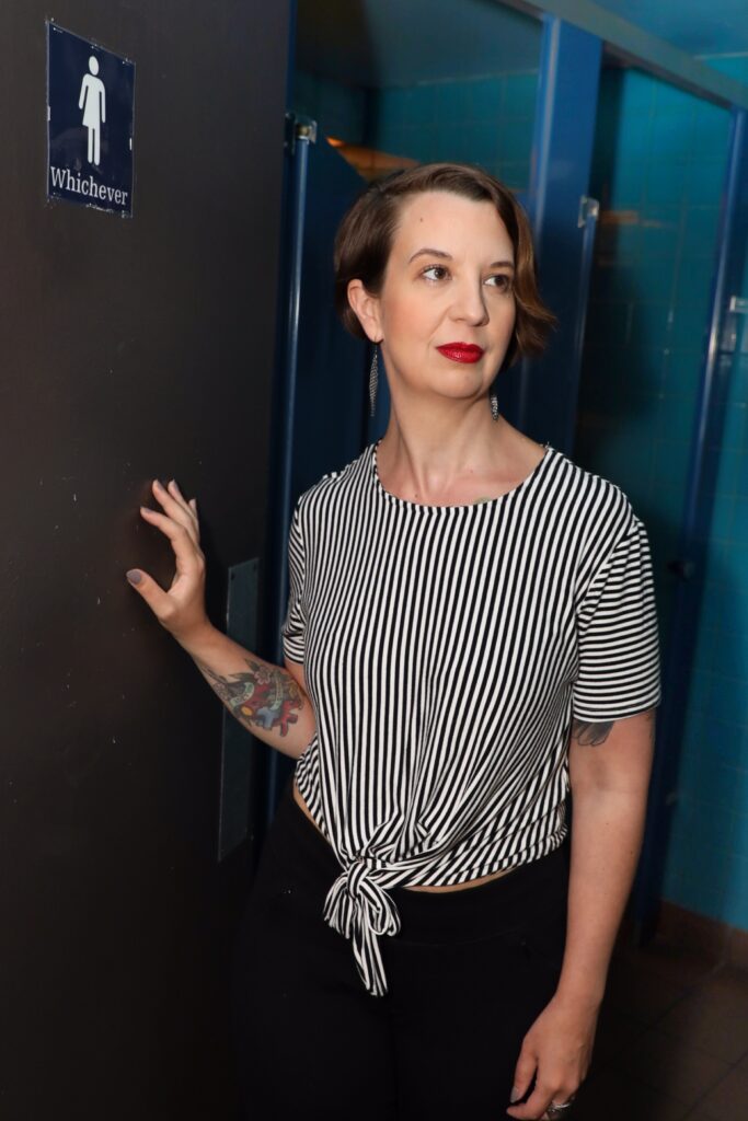 Dr. Leslie Kern standing in a public bathroom next to a sign with a non-binary figure that says "Whichever." Credit: Mitchel Raphael Kern