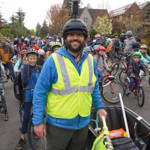 Sam Balto, a young bearded man wearing a bike helmet and reflective vest, stands in front of a bunch of kids with bikes who are part of a bike bus in Portland, Oregon.