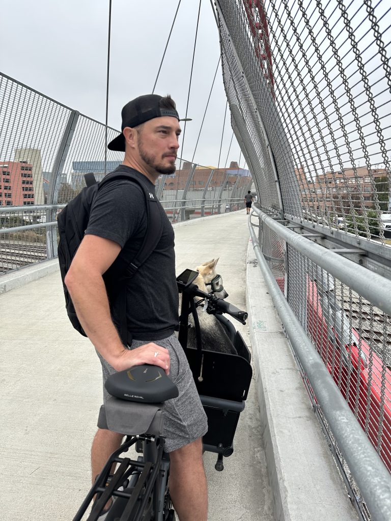 Mayor John Bauters, a man wearing a T-shirt and shorts, stands astride his cargo bike on a bridge, with his big white dog in the bucket.