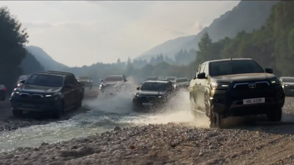 Several large SUVs drive through a riverbed, kicking up water and mud.