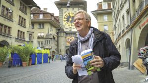 Rick Steves, white man with glasses, wearing brown coat and scarf. He is holding a book and standing in an old European square withe a building with a clock in the background