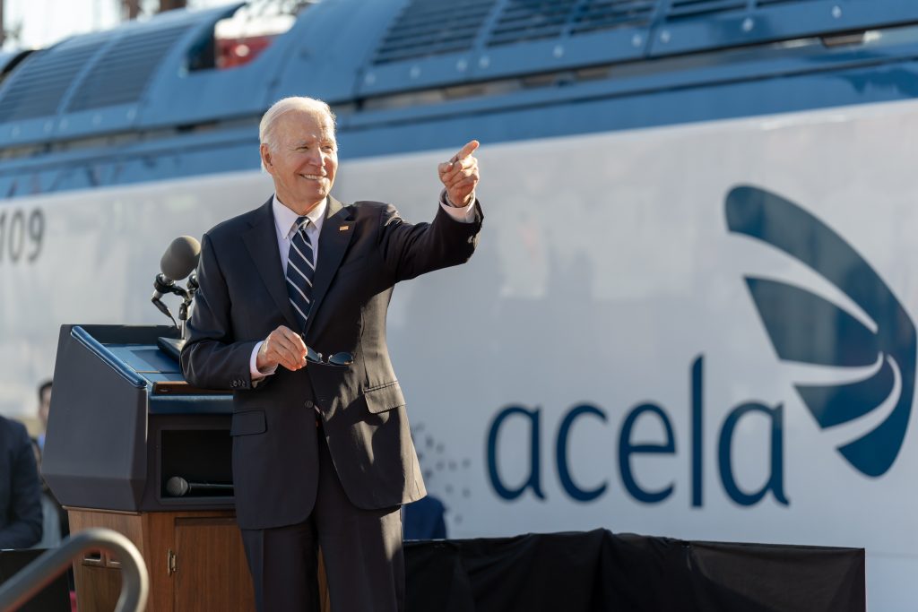 President Joe Biden stands at a podium in front of an Acela train wearing a suit, smiling and pointing