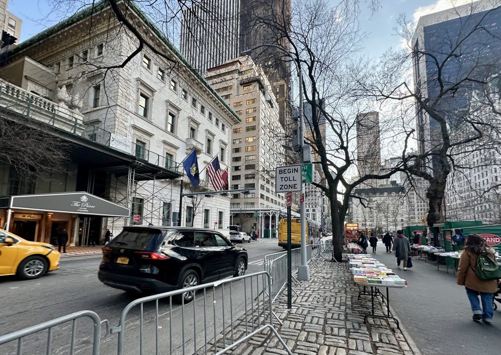 The entrance to the congestion relief zone on Fifth Avenue just north of 60th Street in Manhattan, New York City. A black SUV and a yellow taxi speed past a sign that says "begin toll zone" while a woman walks past a booksellers' tables on the sidewalk.