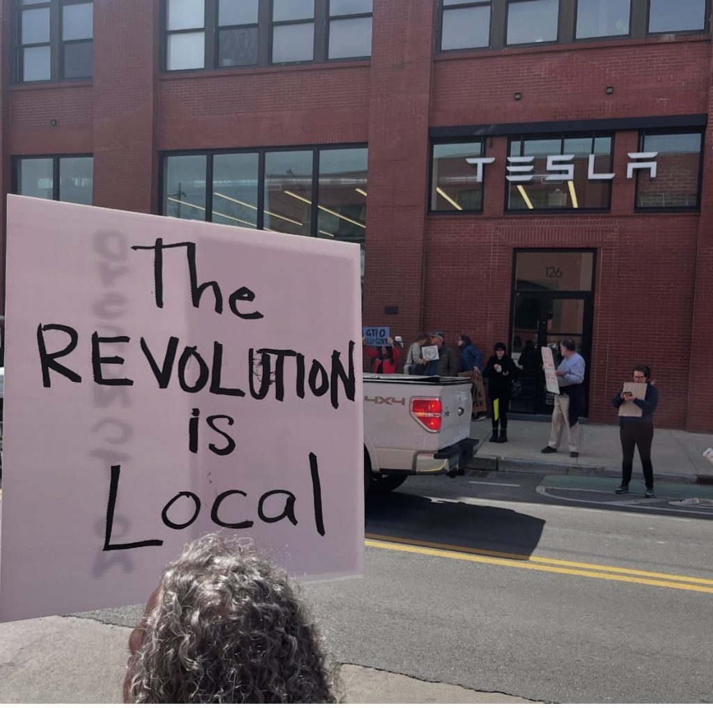 The back of the the head of a protester holding a sign that says "The revolution is local," with a Tesla dealership across the street in the background.