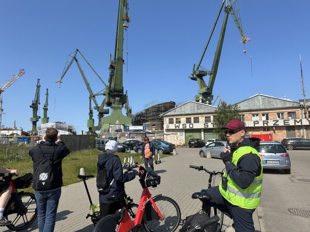People on a bicycle tour of the shipyard in Gdańsk, Poland, stop to take pictures of the shipyard's towering cranes against a bright blue sky while a tour guide explains the history of the site to them.