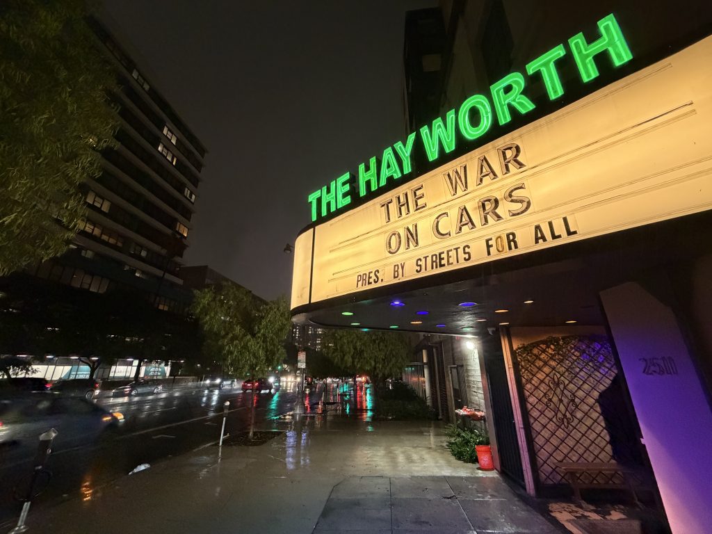 The marquee of the Hayworth Theatre lit up with the theater name in green neon and "The War on Cars Pres. by Streets for All" on display, on a rainy dark night.