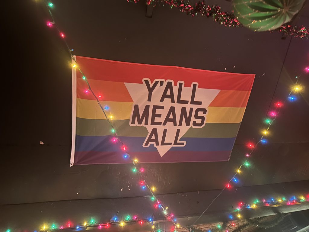 A rainbow flag with the words "Y'ALL MEANS ALL" in black letters on a white upside down triangle, festooned with multicolored lights.