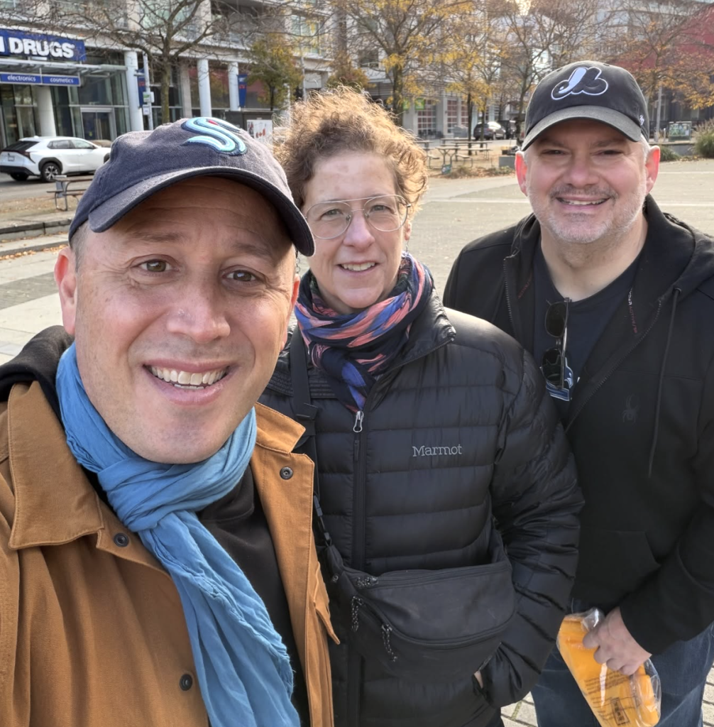 Doug Gordon, wearing a Seattle Kraken hat, Sarah Goodyear, hatless, and Brent Toderian, in a Montreal Expos hat, take a smiling selfie in downtown Vancouver