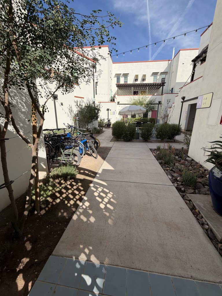A street in the car-free community of Culdesac, in Tempe, Arizona, with white stucco buildings, vegetation, and an ornate shadow cast by a shade screen. 
