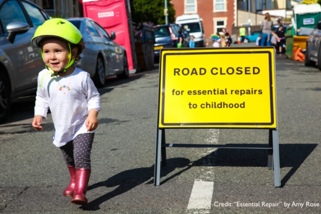 A small girl wearing a bike helmet, maybe three years old, smiles as she walks past a yellow sandwich board on a street that says, "ROAD CLOSED for essential repairs to childhood."