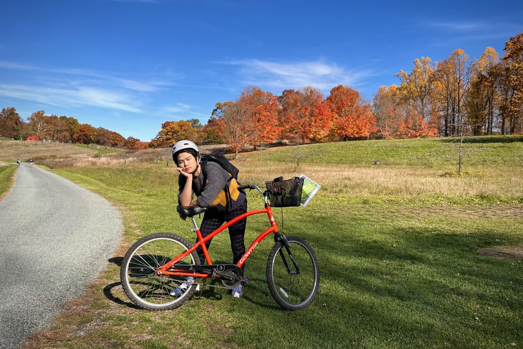 Dr. Rex Tai, a young man wearing a bike helmet and a gray sweater, poses with his red Townie bike in a pastoral autumn landscape. In the distant background, two other cyclists ride on a winding gravel road.
