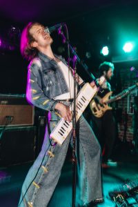 Aaron Oliver, a musical artist known as Pondless, performs on a stage at the Mercury Lounge in New York City, playing keytar, singing into a microphone, and wearing a jean jacket and pants over a white T-shirt