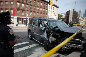 A black SUV on a NYC street with a crumpled hood, obviously the aftermath of a severe crash. The scene is protected by yellow tape, and a woman police officer looks on from the left of the frame.