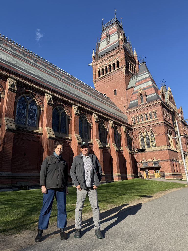 Sarah and Doug stand smiling on the Harvard campus, in front of a massive and elaborate Victorian building.