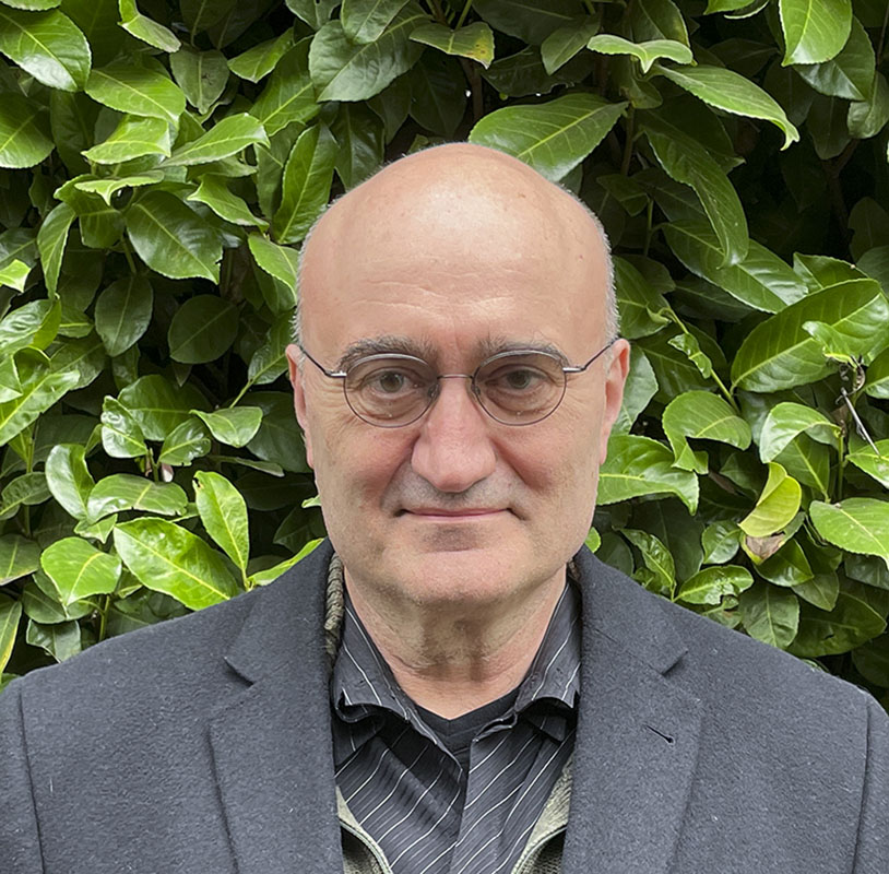 A headshot of Jarrett Walker, wearing glasses and with a smile, standing in front of some greenery