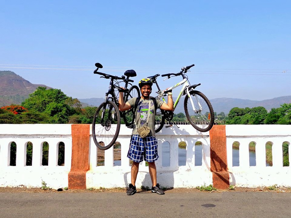 Ashik Jain, a young man wearing shorts and a bicycle helmet, stands in front of a white railing on a bridge with mountains in the background, holding up two bicycles, one in each hand