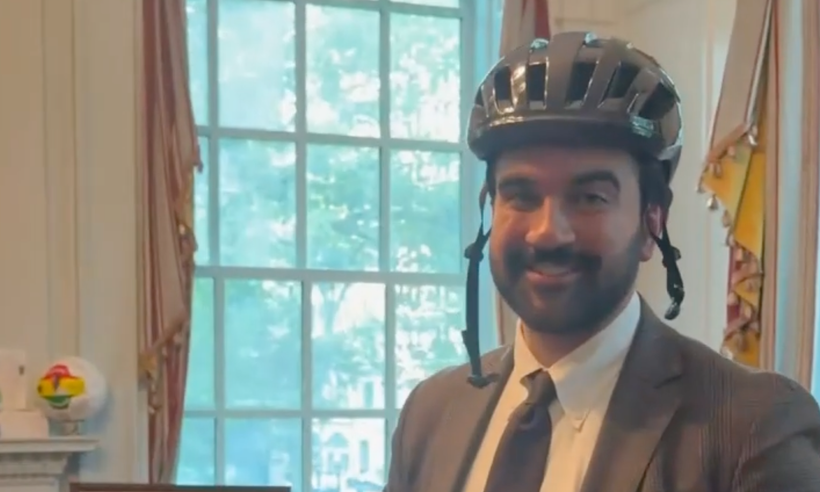 NYC mayor Zohran Mamdani in his office in his trademark suit and tie wearing a bike helmet with the chin strap dangling and a smile on his face.