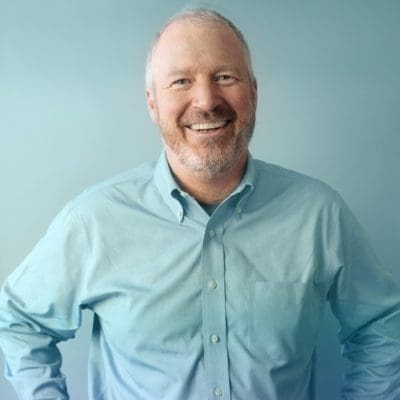 Mike McGinn, a white man wearing a blue shirt with short hair and a salt and pepper beard, stands smiling with hands on hips