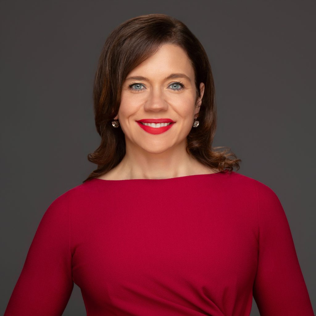 A headshot of Nora Leerhsen, smmiling, with bright red lipstick that matches her dress